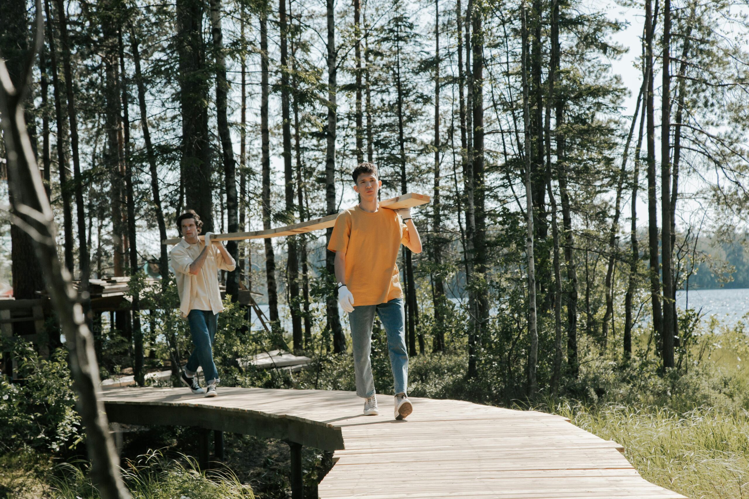 Two young men carrying a plank along a lakeside wooden boardwalk